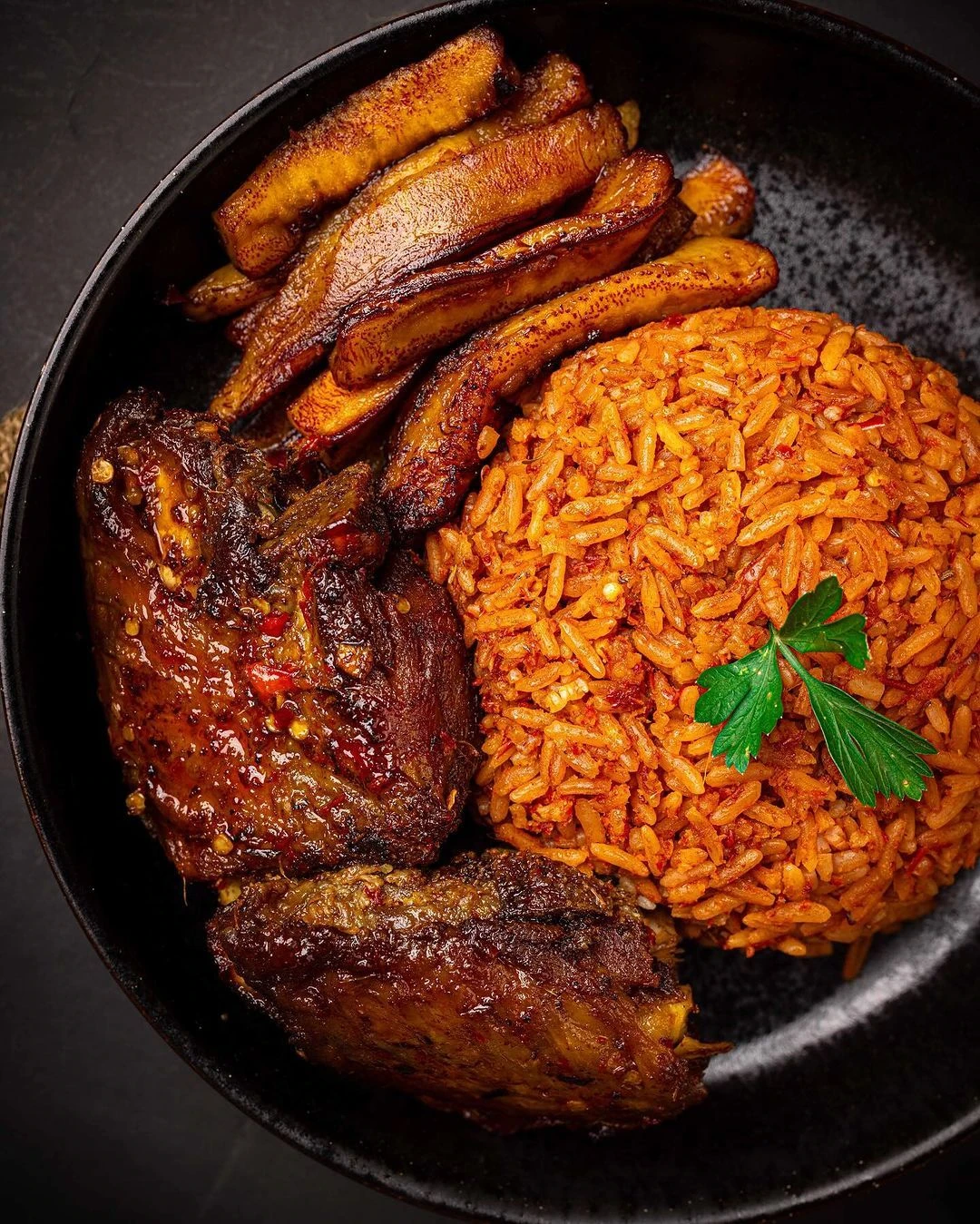 Jollof rice with glazed chicken and fried plantain on a black plate